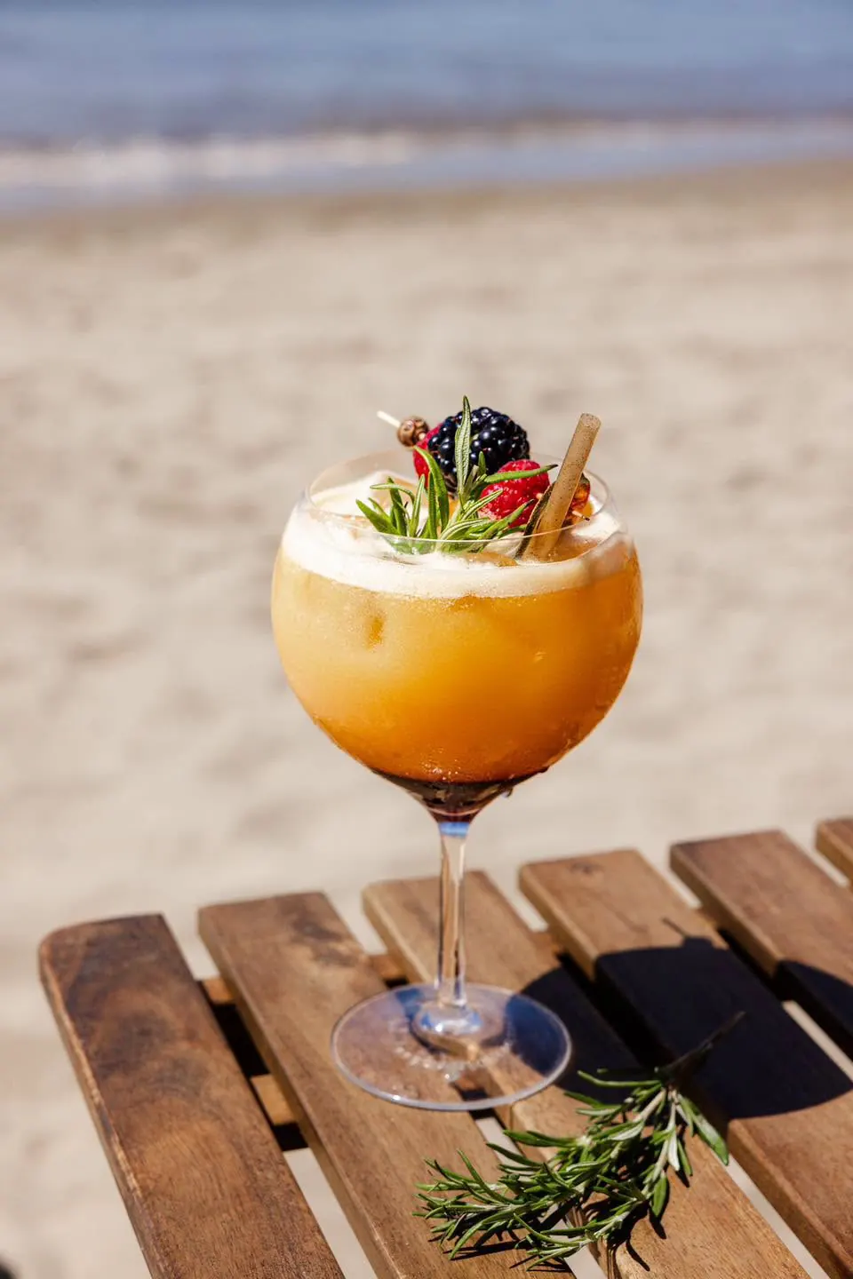 A cocktail garnished with berries and rosemary on a beach table