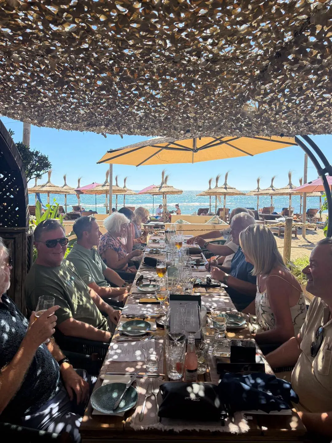 A long table of guests at lunch under the canopy with umbrellas and sea beyond